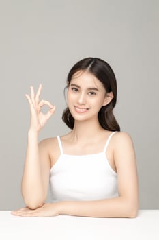 Beautiful young Asian woman model showing OK sign with perfect clean fresh skin on grey background. Face care, Facial treatment, Cosmetology, Plastic Surgery, Lovely girl portrait in studio. photo