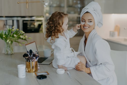 Pretty small girl with curly hair looks how mother does cosmetic procedures applies collagen patches under eyes for skin treatment wear bathrobes pose near desk with beauty products. Everyday care photo
