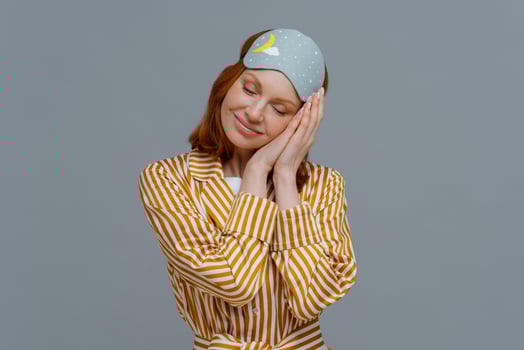 Pleased cheerful woman wears blindfold and striped pajamas, has happy expression, awakes in good mood, has healthy sleep habits, feels totally relaxed, smiles broadly, isolated on grey background photo