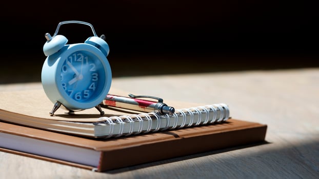 Selective focus at little blue alarm clock with ballpoint pen and kraft paper books with sunlight and shadow on wooden table surface, hobby and relaxing time concept photo
