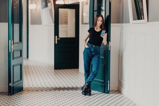 Full length shot of serious fashionable woman concentrated into cell phone, wears black t shirt, jeans and boots, reads information in internet, stands indoor near doors connected to wireless internet photo