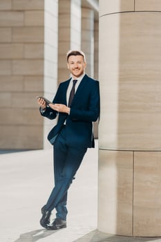 Photo of cheerful male dressed in expensive black suit, stands crossed legs, works on touch pad, stands in modern office interior, smiles positively at camera. Young male manager. Full length portrait