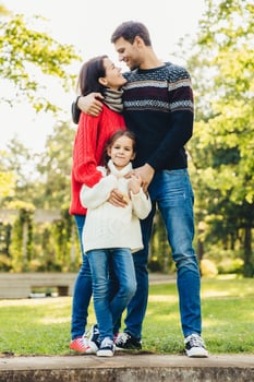 Young happy family of three stand together outdoors, embrace, have good relationships. Couple in love look at each other, hug their little daughter. Happiness and harmony in family life. photo