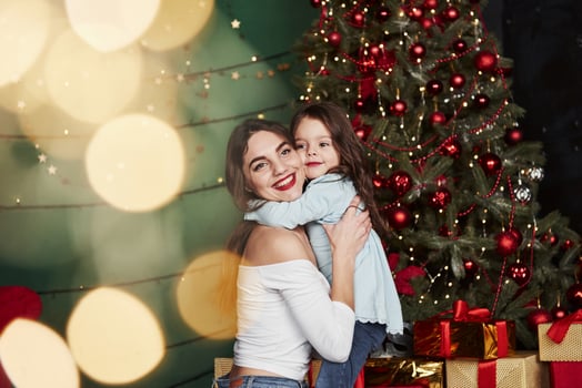 Many of gift boxes on the floor. Cheerful mother and daughter hugging each other near the Christmas tree that behind. Cute portrait photo