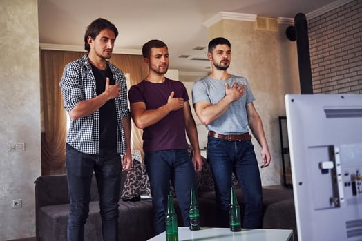Three friends stands indoors against TV with hands on heart. National hymn playing photo