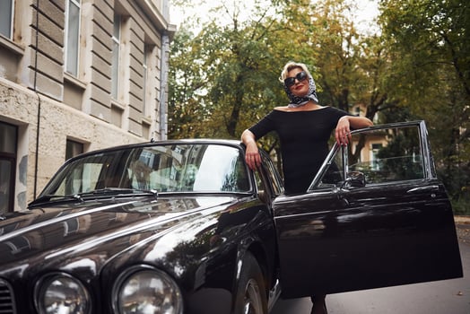 Blonde woman in sunglasses and in black dress near old vintage classic car photo