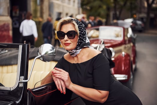 Blonde woman in sunglasses and in black dress leaning on old vintage classic car photo