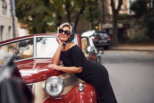 Blonde woman in sunglasses and in black dress leaning on old vintage classic car photo