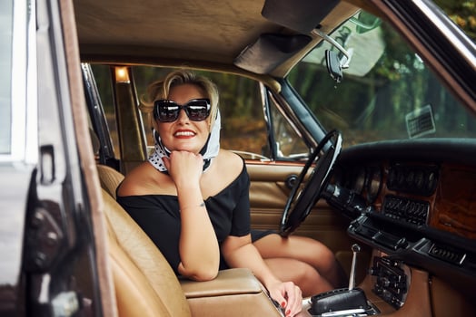 Blonde woman in sunglasses and in black dress sits in old vintage classic car photo