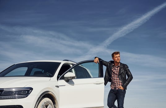 Man in black leather jacket stands near his parked white car outdoors against blue sky photo