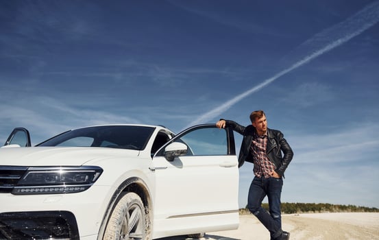 Man in black leather jacket stands near his parked white car outdoors against blue sky photo