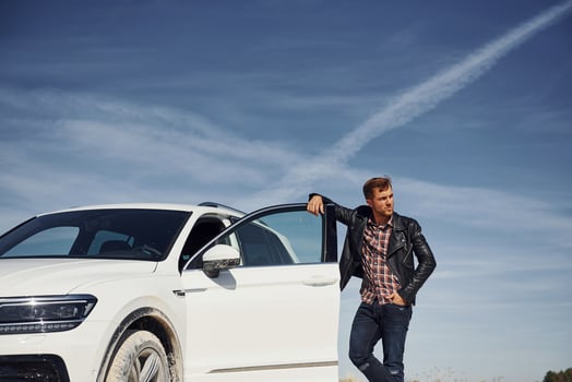 Man in black leather jacket stands near his parked white car outdoors against blue sky photo