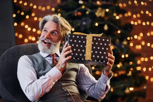 Portrait of stylish senior with grey hair and beard is in decorated christmas room with gift box photo
