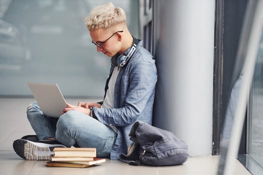 Young hipster guy sitting indoors near grey background with laptop and books photo