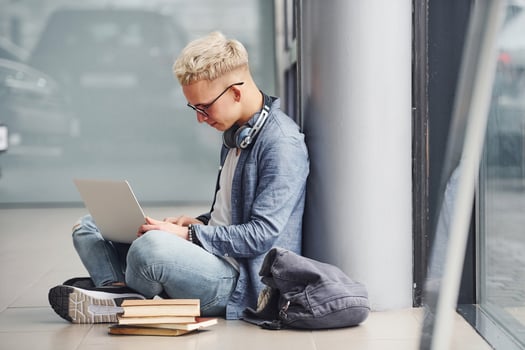 Young hipster guy sitting indoors near grey background with laptop and books photo