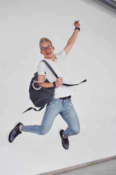 Happy hipster guy in casual clothes and with backpack jumping indoors against white wall photo
