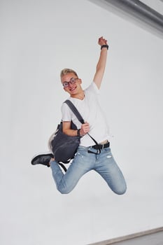 Happy hipster guy in casual clothes and with backpack jumping indoors against white wall photo