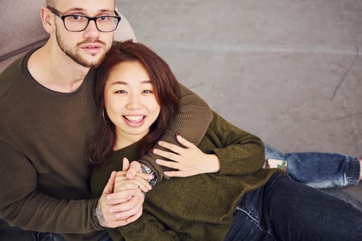 Happy multi ethnic couple in casual clothes sitting together indoors in the studio. Caucasian guy with asian girlfriend photo