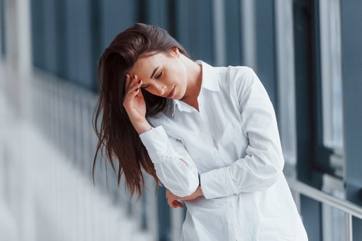 Brunette in white shirt standing indoors in modern airport or hallway at daytime and having headache photo