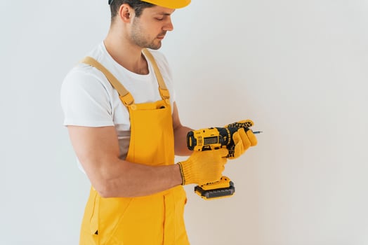 Handyman in yellow uniform standing against white wall with automatic screwdriver. House renovation conception photo