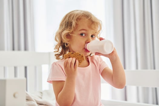 Sitting on the bed, eating cookies and drinking milk. Cute little girl in casual clothes is indoors at home at daytime photo