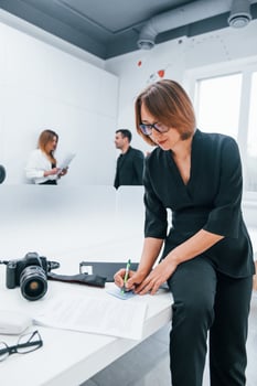 Woman in black formal clothes and with camera in the office. Colleagues behind photo