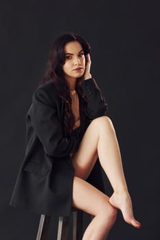Sexy brunette with curly hair in luxury clothes sits on chair in the studio against black background photo