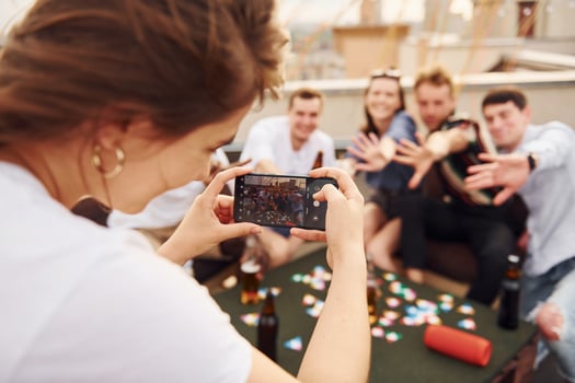 Girl doing photo when people playing card game. Group of young people in casual clothes have a party at rooftop together at daytime
