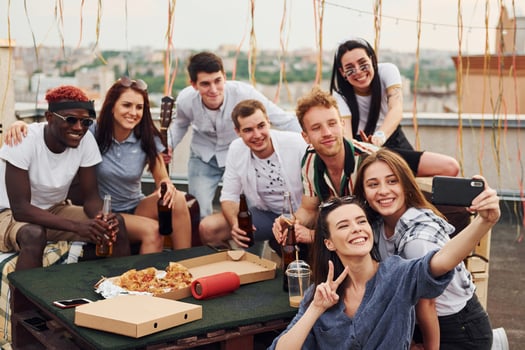 Girl making selfie. With delicious pizza. Group of young people in casual clothes have a party at rooftop together at daytime photo