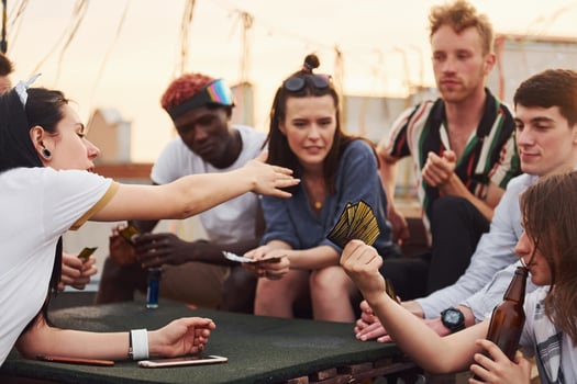 Sitting by table and playing card game. Group of young people in casual clothes have a party at rooftop together at daytime photo