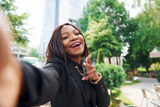 Doing selfie. Young afro american woman in fashionable clothes outdoors in the city near green trees and against business building photo