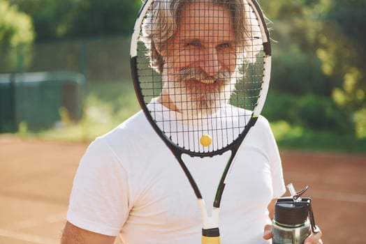 Taking a break and drinking water. Senior modern stylish man with racket outdoors on tennis court at daytime photo