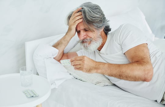 Sick senior stylish modern man with grey hair and beard indoors lying on the bed and feels bad photo