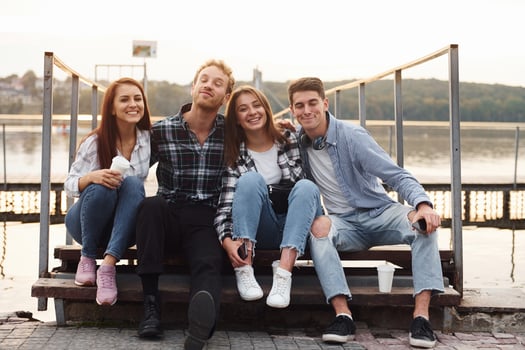 Sitting near lake and embracing each other. Group of young cheerful friends that is outdoors having fun together photo