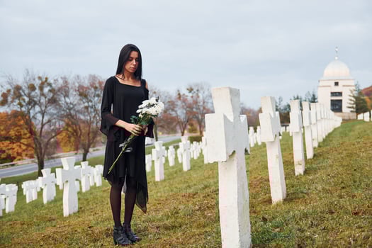 With flowers in hands. Young woman in black clothes visiting cemetery with many white crosses. Conception of funeral and death photo
