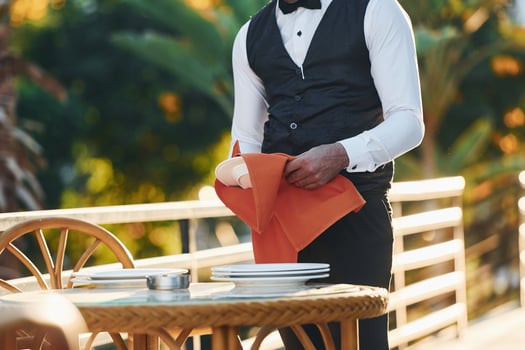 Wipes plates by red cloth. Black waiter in formal clothes is at his work outdoors at sunny daytime photo