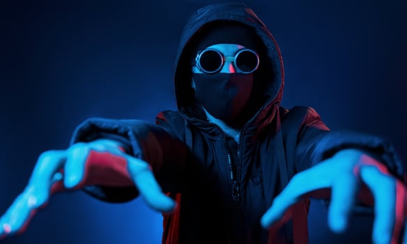 In mask, eyewear and hood. Neon lighting. Young european man is in the dark studio photo
