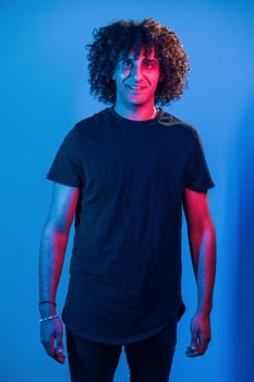 Posing for a camera. Young beautiful man with curly hair is indoors in the studio with neon lighting photo