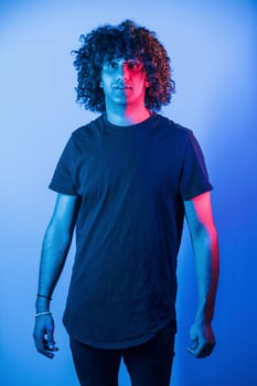 Posing for a camera. Young beautiful man with curly hair is indoors in the studio with neon lighting photo