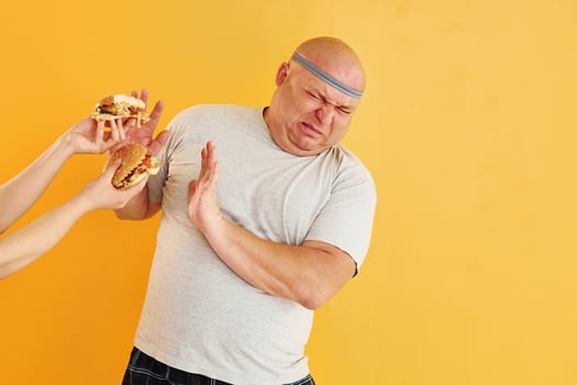 Hands with hamburgers temptates guy. Funny overweight man in sportive head tie is against yellow background photo