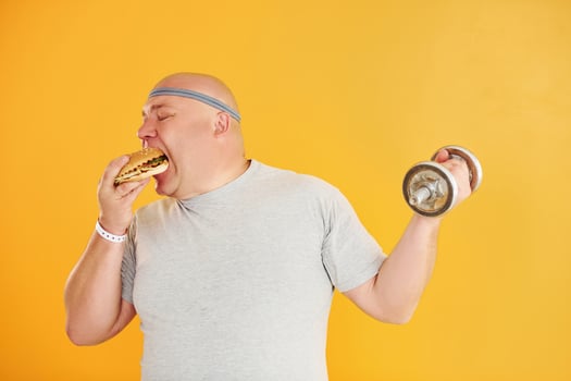 Holds hamburger and dumbbell. Funny overweight man in sportive head tie is against yellow background photo