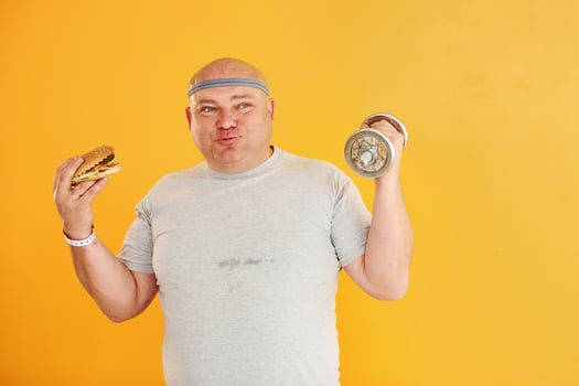 Holds hamburger and dumbbell. Funny overweight man in sportive head tie is against yellow background photo