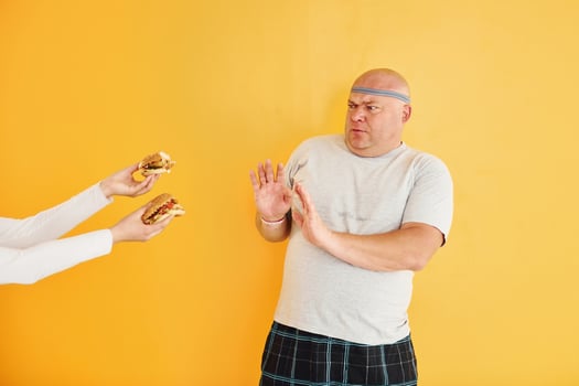 Hands with hamburgers temptates guy. Funny overweight man in sportive head tie is against yellow background photo