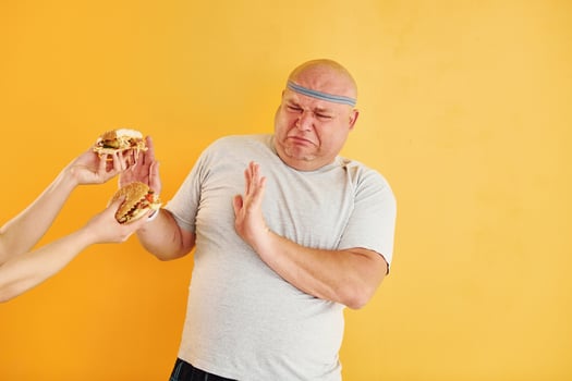 Hands with hamburgers temptates guy. Funny overweight man in sportive head tie is against yellow background photo