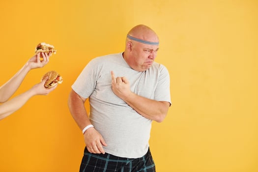 Hands with hamburgers temptates guy. Funny overweight man in sportive head tie is against yellow background photo