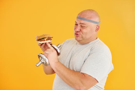 Holds hamburger and dumbbell. Funny overweight man in sportive head tie is against yellow background photo