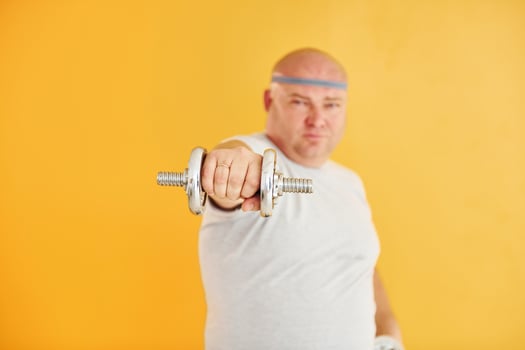 Exercises by using dumbbells. Funny overweight man in sportive head tie is against yellow background photo