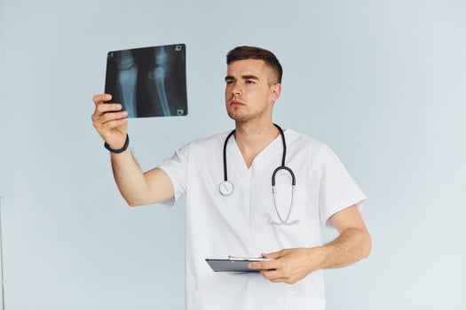 With stethoscope. Young male doctor in uniform is indoors against white background photo