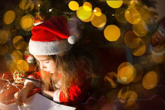Little girl in a Santa hat and red dress under Christmas tree is dreaming, waiting for the holiday, lying on a plaid blanket. A letter on piece of paper, gifts. New year, Christmas. defocus lights photo