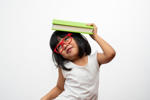 Funny and Happy Asian little preschool girl wearing red glasses holding a green book on the head, on white isolated background. Concept of school kid and education in elementary and preschool photo